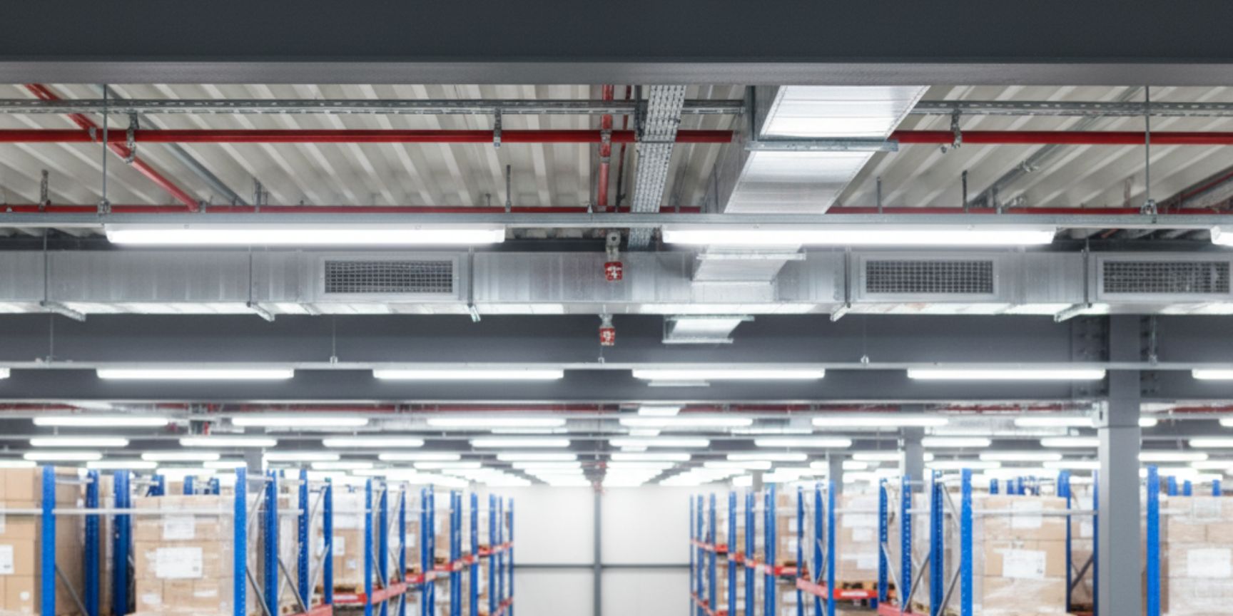 Detailed view of the ceiling construction of a logistics hall, taken from the perspective of a high shelf. In the foreground, a massive grey truss of the roof construction can be seen, which represents the